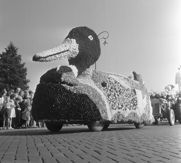 Ein Schwarz-Weiß-Foto eines Entenwagens bei einem Umzug, geschmückt mit Blumen, mit einer Gruppe von Menschen auf der linken Seite und Bäumen und einem klaren Himmel im Hintergrund.