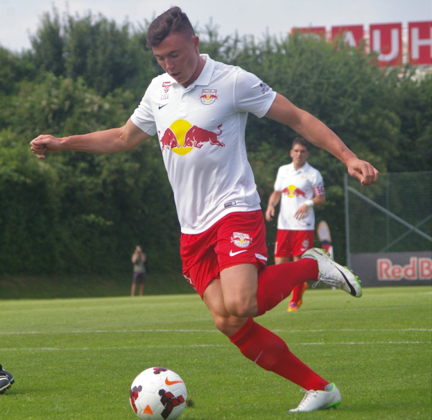 Ein Mann in einem Fussballtrikot schießt einen Ball auf einem grünen Rasen mit Bäumen und einem klaren blauen Himmel im Hintergrund, wobei ein Schild mit der Aufschrift "RB Leipzig" sichtbar ist.