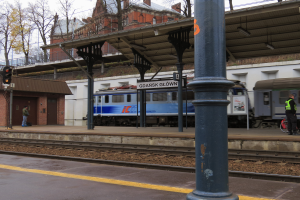 Ein Zug fährt auf einen Bahnsteig zu, auf dem eine Person mit einem Fahrrad steht, mit Schienen, Lichtern, Schildern, einer überführung, Bäumen, Gebäuden und Himmel im Hintergrund.