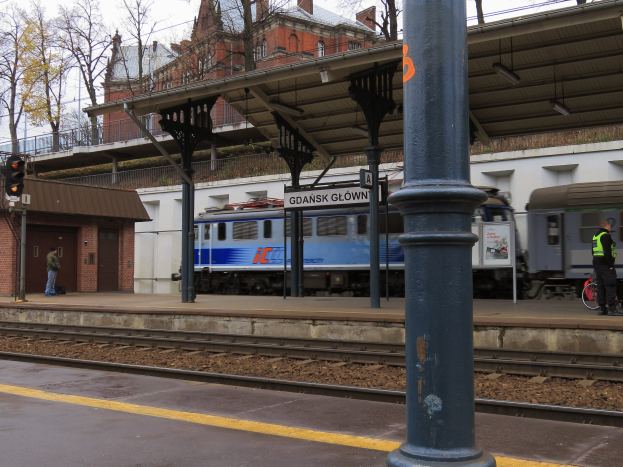 Ein Zug fährt auf einen Bahnsteig zu, auf dem eine Person mit einem Fahrrad steht, mit Schienen, Lichtern, Schildern, einer überführung, Bäumen, Gebäuden und Himmel im Hintergrund.