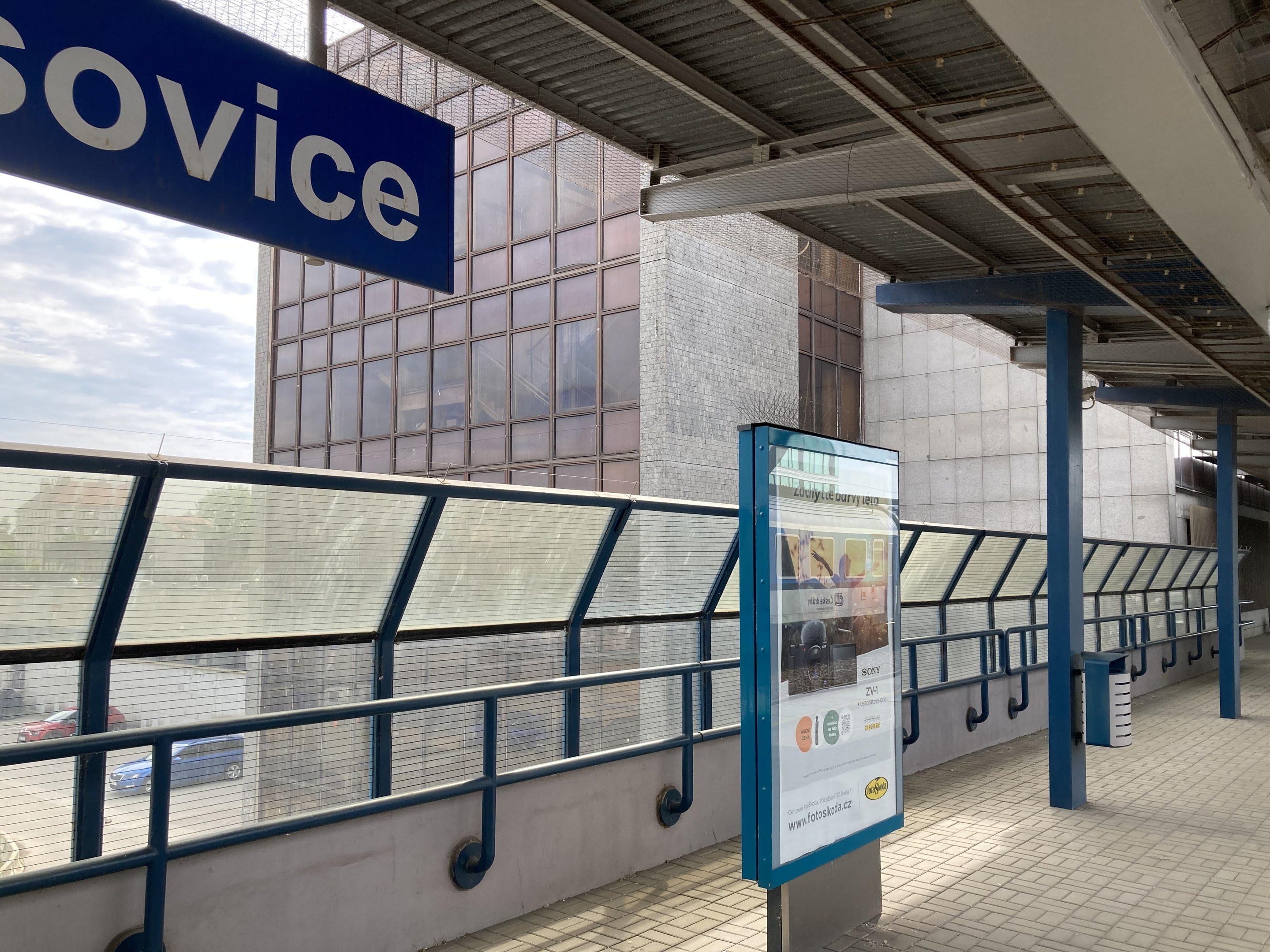 Haltestelle mit einem "Service"-Schild, Infotafel, Metallzaun, Masten, Dach mit Deckenleuchten, Gebäude mit Fenstern, Fahrzeuge auf der Straße, Bäume und ein bewölkter Himmel.