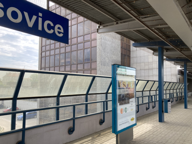 Haltestelle mit einem "Service"-Schild, Infotafel, Metallzaun, Masten, Dach mit Deckenleuchten, Gebäude mit Fenstern, Fahrzeuge auf der Straße, Bäume und ein bewölkter Himmel.