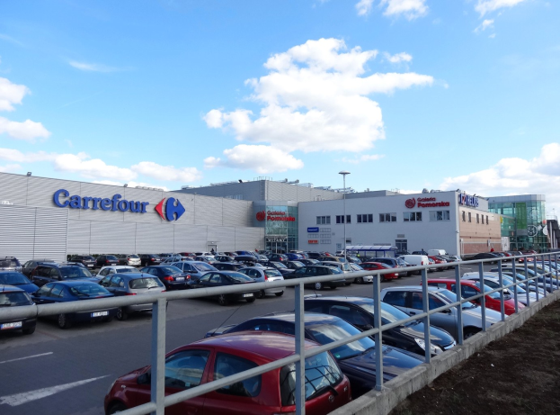 Großer Autohandel mit parkenden Fahrzeugen, umgeben von städtischer Infrastruktur wie Gebäuden, Straßenlaternen und Fahrzeugen auf der Straße.
