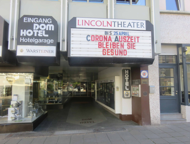 Außenansicht des Lincoln Theaters in Berlin, Deutschland, mit Glasfenstern und -türen und einer Tafel mit Text sowie einem Innenraum, der eine pulsierende Stadtlandschaft suggeriert.