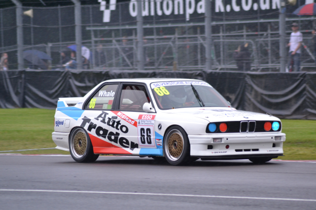 Ein weißer Rennwagen fährt auf einer Strecke mit Zuschauern hinter einem Maschendrahtzaun, einige halten Schirme, und ein Banner im Hintergrund.