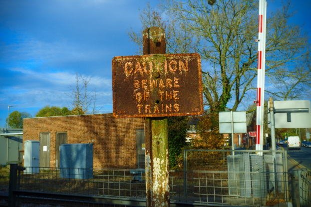 Vorsichtsschild an einem Bahnübergang-Zaun, mit Bäumen, Strommasten, einem Gebäude, Containern, Fahrzeugen und einem bewölkten Himmel.