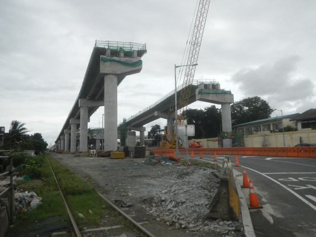 Eine Baustelle mit einer Brücke im Hintergrund, eine Straße mit Absperrgittern auf der rechten Seite, Steine und Gras am Boden, eine Eisenbahnschiene auf der linken Seite, Bäume und Gebäude auf beiden Seiten der Straße und ein bewölkter Himmel.