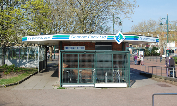 Bus-Haltestelle mit 'Gosport Ferry Ltd'-Schild, Metallzaun, Menschen, Fahrrädern, Bäumen, Straßenlaternen, Schildern, einem Gebäude und einem bewölkten Himmel.