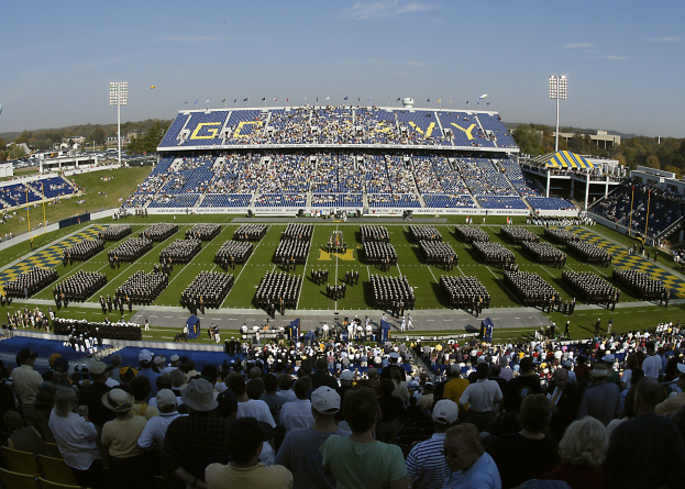 Eine große Menschenmenge schaut einem Fußballspiel in einem grünen Stadion unter einem klaren blauen Himmel zu, mit Laternen, Bäumen und Gebäuden im Hintergrund.