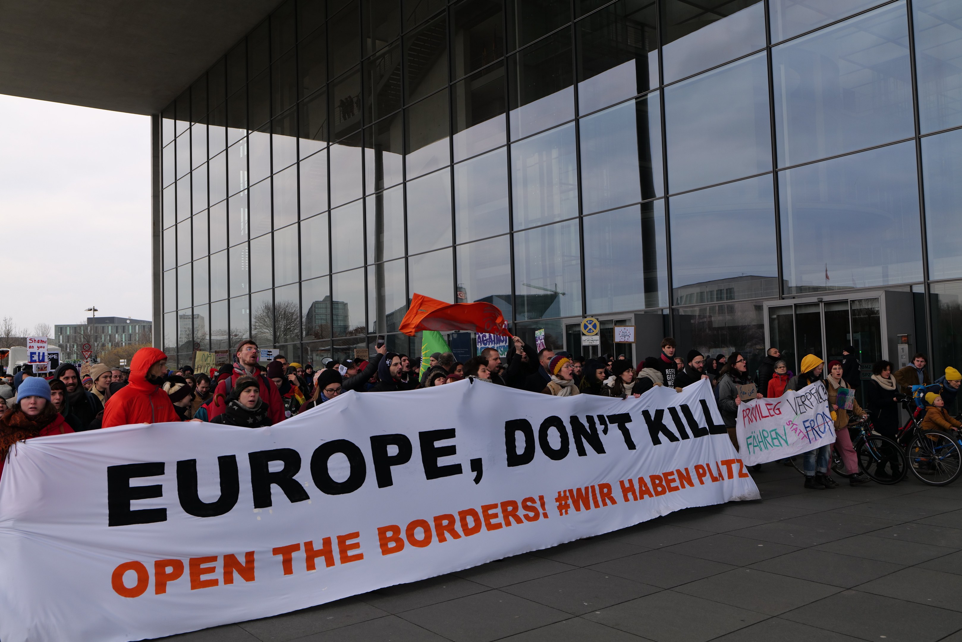 Gruppe von Menschen mit einem Transparent "Europa, töte nicht, öffne die Grenzen" vor einem Glasgebäude, einige tragen Mützen und halten Fahnen oder Fahrräder, mit Bäumen und einem klaren blauen Himmel im Hintergrund.
