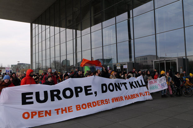 Gruppe von Menschen mit einem Transparent "Europa, töte nicht, öffne die Grenzen" vor einem Glasgebäude, einige tragen Mützen und halten Fahnen oder Fahrräder, mit Bäumen und einem klaren blauen Himmel im Hintergrund.