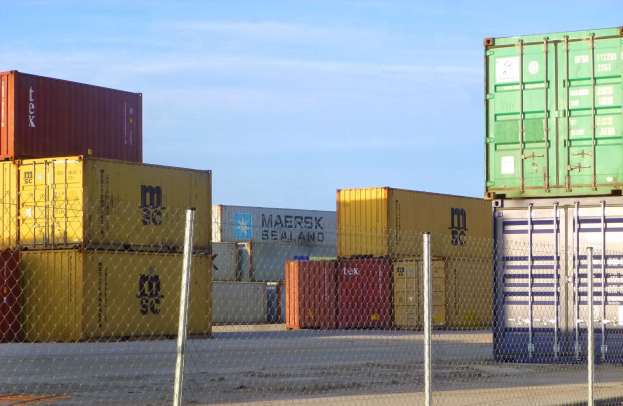 Ein hoher Stapel von Containern hinter Maschendrahtzaun, mit einer Straße unten und bewölktem Himmel im Hintergrund.