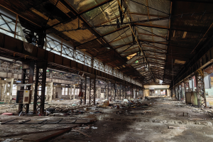 Ein verlassenes Fabrikinnere mit verstreuter Trümmern, zerbrochenen Fenstern, Metallstangen, Deckenleuchten und einem beschädigten Dach.