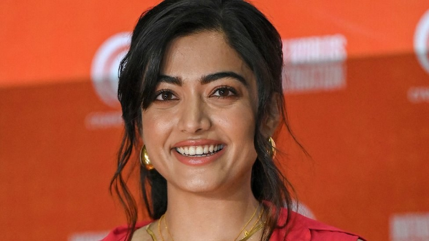 Eine Frau in einem roten Kleid mit einem eleganten Dutt und natürlichem Make-up lächelt in die Kamera, trägt schwarze High Heels und hat einen hellgelben Banner mit der Aufschrift 'Die besten Schönheits Looks von Sonam Kapoor beim Cannes Film Festival' im Hintergrund.