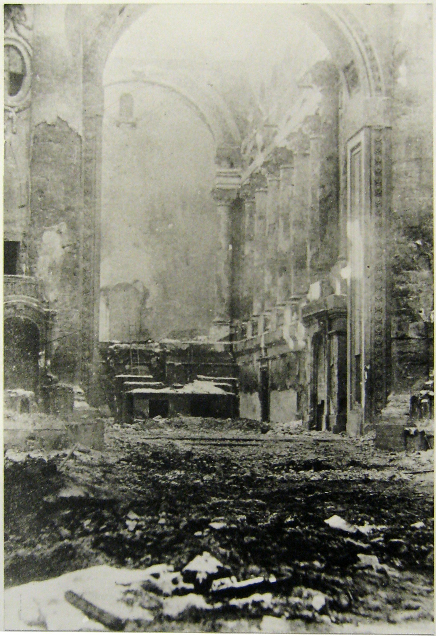 Schwarzes und weißes Foto eines feuerbeschädigten Gebäudes mit Trümmern auf dem Boden und sichtbaren Wänden und Säulen mit Text unten.