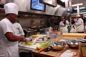 Eine Gruppe von Köchen in einer Küche bei der Zubereitung von Essen auf einem Schneidbrett, mit einem Koch im Vordergrund in einem weißen Kleid und einer Mütze, der ein Messer hält, und umgeben von Utensilien, Schüsseln, Gemüse, einem Mobiltelefon, Flaschen und anderen Gegenständen, mit einem Fernseher und Deckenlampen im Hintergrund.