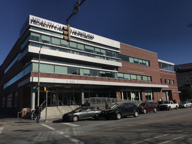 Großes Gebäude mit der Aufschrift "New York City Health Care Homeless Center" mit parkenden Autos, Passanten, Straßenmöblierung und Bäumen unter einem klaren Himmel.