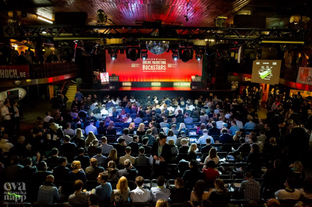 Eine große Menschenmenge sitzt auf Stühlen vor einer Bühne mit Bildschirmen und Bannern, beleuchtet von Scheinwerfern und umgeben von Geländern, mit einem Wasserzeichen in der linken unteren Ecke, in Erwartung eines Online-Marketing-Rockstars-Events.