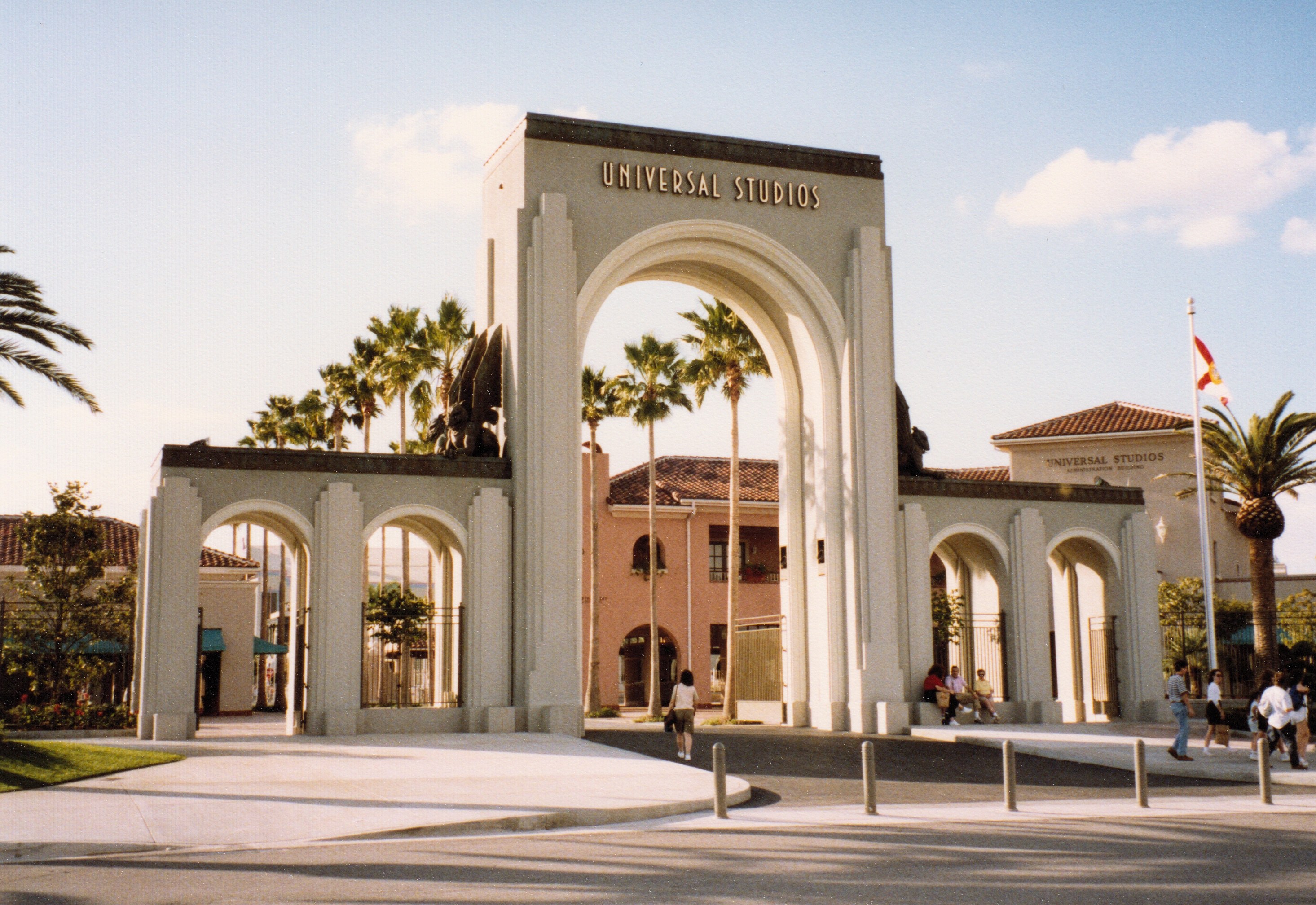 Eingang von Universal Studios Hollywood mit einem Bogen, einer Statue, Menschen, Bäumen, Gebäuden, einer Fahne und einem bewölkten Himmel.