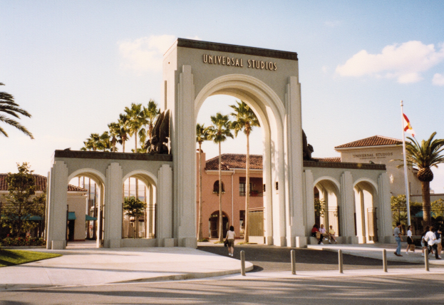 Eingang von Universal Studios Hollywood mit einem Bogen, einer Statue, Menschen, Bäumen, Gebäuden, einer Fahne und einem bewölkten Himmel.