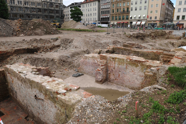 Römische Ruinen umgeben von modernen städtischen Elementen einschließlich Gebäuden, Bäumen, Fahrzeugen und Menschen.