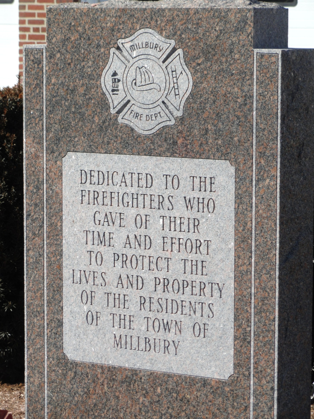 Gedenkstein mit einer Gedenktafel für Feuerwehrleute in Millbury, aufgestellt vor einem Backsteingebäude mit umliegender Vegetation.