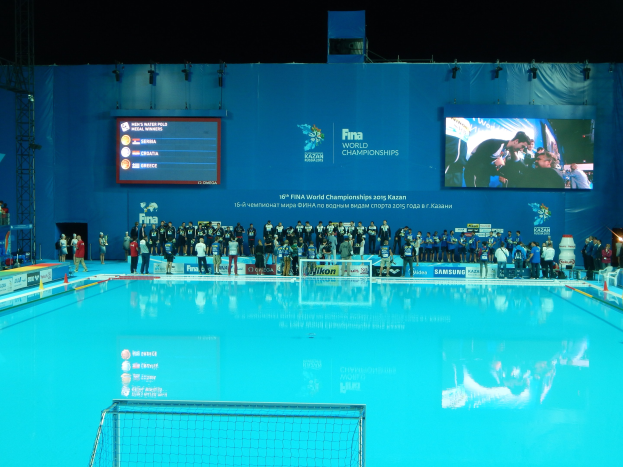 Eine Gruppe von Menschen um einen Hallenbadpool mit Netz am Boden herumstehend, mit einer Wand mit 'FINA World Championships 2015 Kazan' Text und Logos, Trägern und Lichtern im Hintergrund.