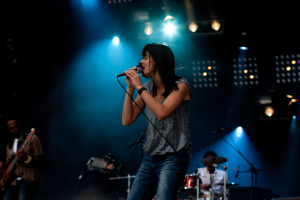 Eine Frau singt in ein Mikrofon auf der Bühne eines Konzerts, flankiert von einem Mann, der Gitarre spielt, und einer Person, die Schlagzeug spielt, unter hellen Bühnenlichtern.