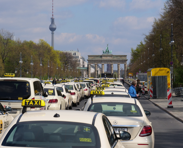Eine belebte Straße in Berlin mit zahlreichen parkenden Taxis, Fußgängern auf dem Gehweg, Laternen, Bäumen, Gebäuden, einem fernen Bogen mit Statuen und einem Turm sowie einem bewölkten Himmel.