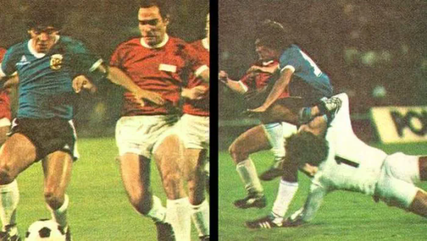 Eine Gruppe von Männern, die Fußball auf einem Feld spielen, mit einem, der den Ball tritt, und anderen, die versuchen, die Kontrolle zu übernehmen, und einer Tafel im Hintergrund, die eine Stadionatmosphäre suggeriert.