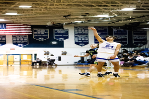 Ein Mann in Sportbekleidung schießt einen Basketball in einer Turnhalle mit Zuschauern auf Tribünen im Hintergrund.
