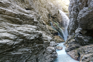 Kleiner Wasserfall, der über zerklüftete Felsen in einem steinigen Tal hinabfließt, umgeben von saftig grünen Hügeln, mit kristallklarem Wasser und hellem Sonnenlicht, das die Szene erhellt.