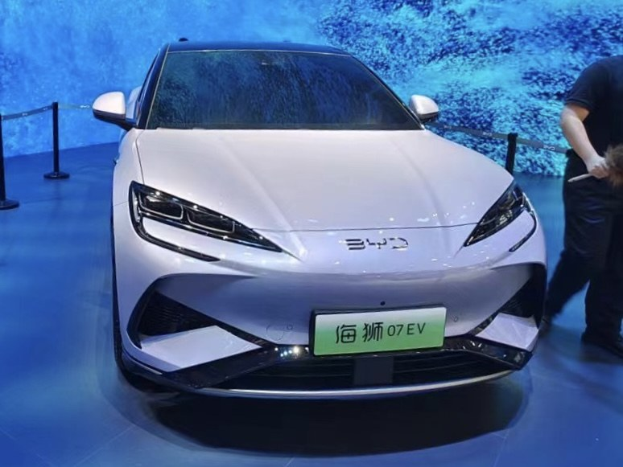 Weißer Wagen auf einer Autauschau mit einer Person, die rechts davon ein Objekt hält, einem Bildschirm im Hintergrund und Absperrpollern mit Seilen links.