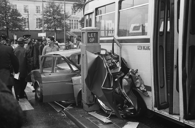 Schwarz-weißes Bild eines Busses, der auf der Seite der Straße in ein Auto gekracht ist, mit mehreren Menschen, die um die Szene stehen, einige mit Mützen, und Bäumen und Gebäuden mit Fenstern im Hintergrund.
