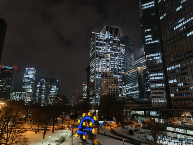 Europäische Zentralbank-Hauptsitz in Frankfurt, Deutschland nachts mit beleuchteten Gebäuden, Bäumen, Straßenlaternen, Lichtern und Fußgängern unter einem bewölkten Himmel.