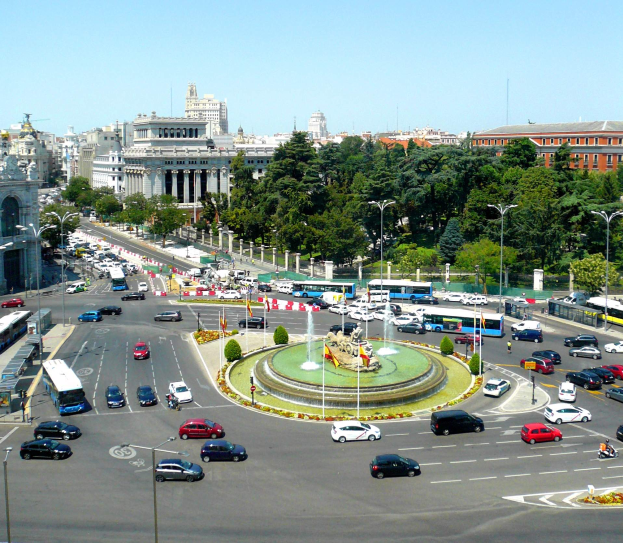Eine belebte Stadtstraße mit verschiedenen Fahrzeugen, einem zentralen Brunnen, Laternen, Bäumen, Gebäuden und einem klaren blauen Himmel.
