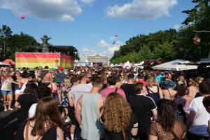 Eine große Menschenmenge, die eine Straße entlanggeht, die mit Zelten, Bäumen, Pfählen, Lichtern und einer Statue gesäumt ist, während des Berliner Pride-Festivals, mit Gebäuden und einem Himmel voller Wolken und Ballons im Hintergrund.