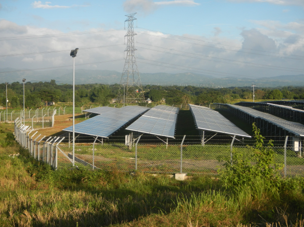 Ein Solarpanelfeld, umgeben von einem Zaun, mit Gras, Pflanzen und Bäumen und einem Übertragungsturm mit Drähten im Hintergrund bei einem bewölkten Himmel.