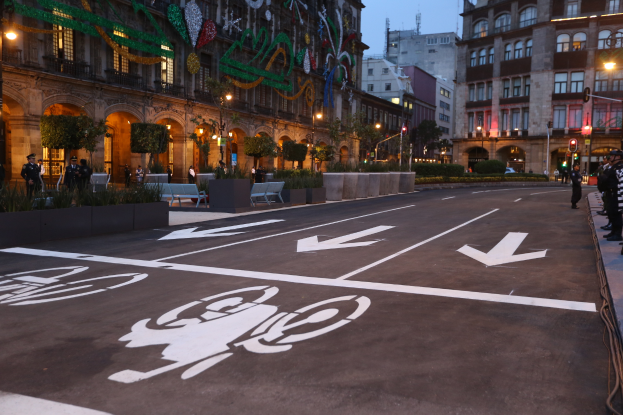 Eine Stadtstraße mit einem Fahrradweg, Gebäuden, Straßenlaternen, Ampeln, Menschen auf dem Gehweg, Bänken, Pflanzen, Bäumen, dekorativen Elementen und einem klaren blauen Himmel.