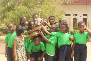 Gruppe von Kindern mit einem Pokal in einem Schulhof mit Bäumen und einem Gebäude im Hintergrund.