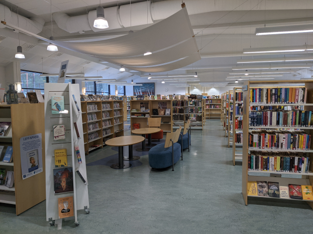 Eine geräumige Bibliothek mit zahlreichen Büchern auf Regalen, Tischen und Stühlen, Sofas, Texttafeln, Lampen, Rohren und anderen Gegenständen, mit Glasfenstern im Hintergrund, die eine Bibliothek-Atmosphäre schaffen.