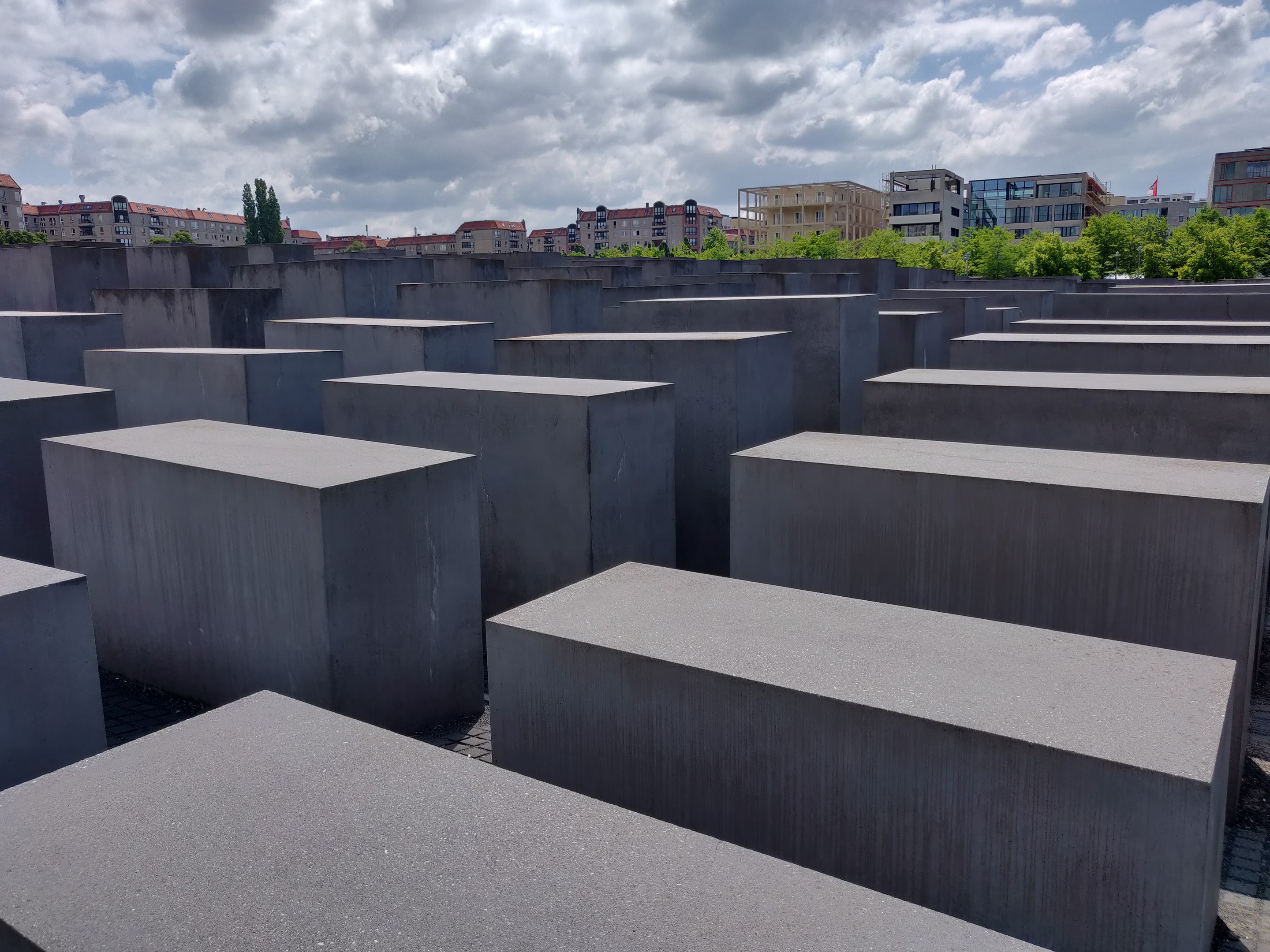 Konkreter Holocaust-Mahnmal für die ermordeten Juden Europas in Berlin, Deutschland, mit Bäumen, Gebäuden und Wolken im Hintergrund.