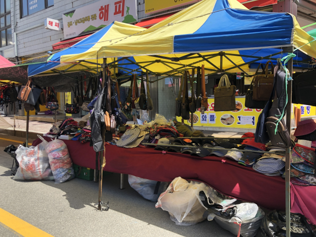 Ein Straßenmarkt mit Zelten, die Taschen, Hüte und andere Gegenstände ausstellen, vor Gebäuden mit Fenstern und Texttafeln im Hintergrund.