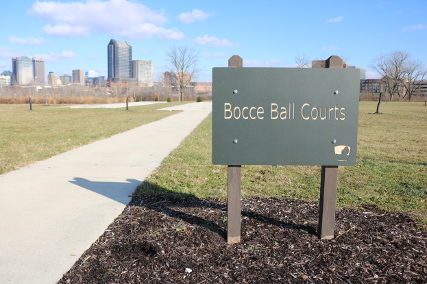 Platte mit Text "Bocce Ball Courts in Indianapolis, Indiana" auf Gras neben einem Weg, Bäumen, Pfosten, einem Zaun, Gebäuden und einem bewölkten Himmel.