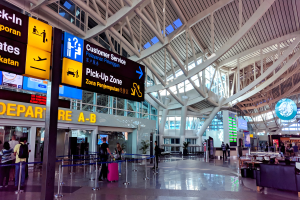 Ein Flughafen-Terminal-Innenraum mit Menschen, die gehen, einige tragen Taschen, und Pfosten mit Bändern und Texttafeln; Sofas und Tische mit Gegenständen auf der rechten Seite und ein gläsernes Gebäude mit Deckenbeleuchtung im Hintergrund.