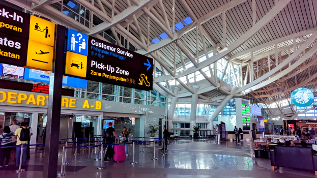 Ein Flughafen-Terminal-Innenraum mit Menschen, die gehen, einige tragen Taschen, und Pfosten mit Bändern und Texttafeln; Sofas und Tische mit Gegenständen auf der rechten Seite und ein gläsernes Gebäude mit Deckenbeleuchtung im Hintergrund.