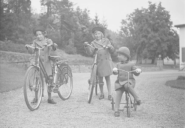 Drei Kinder in Mänteln und Hüten fahren Fahrräder in einem Park mit Bäumen und einem Haus im Hintergrund, in Schwarz-Weiß.
