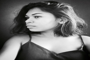 Eine selbstbewusste Frau in einem schwarzen Kleid posiert in einem Schwarz-Weiß-Foto, ihr glatter Bob und das natürliche Make-up unterstreichen ihre selbstbewusste Haltung vor einer neutralen Wand.