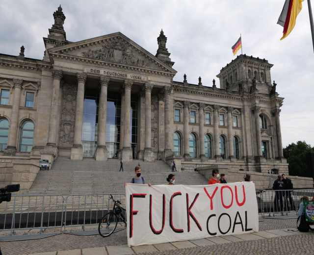 Protestierende halten ein "Fuck You Coal"-Schild vor dem Reichstaggebäude in Berlin, mit Bäumen, einer Flagge und einem bewölkten Himmel im Hintergrund.