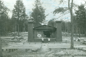 Ein altes Schwarz-Weiß-Foto einer Bühne im Wald mit zwei Personen davor, umgeben von Holzstämmen und Bäumen, mit dem Himmel im Hintergrund.
